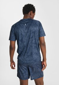 Man i marinblå mönstrad sporttröja och shorts, som står med ryggen mot och ena handen lätt upplyft, mot en enfärgad ljus bakgrund.