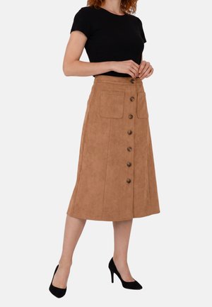 Donna che indossa una maglietta nera a maniche corte, una gonna midi color beige con bottoni davanti e tasche, e scarpe nere con tacchi alti, in piedi davanti a uno sfondo semplice.