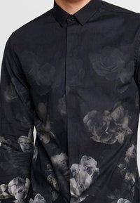 Maglietta nera con una stampa floreale di rose grigie, maniche lunghe, colletto a punta e chiusura con bottoni. Texture liscia.