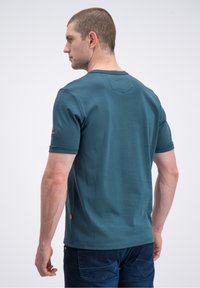 Žalsvai mėlynas trumpomis rankovėmis marškinėliai iš lygaus medvilnės audinio, su V formos nugaros dizainu ir mažu logotipo pleistru kairėje rankovėje.