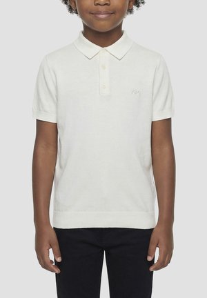 Kind draagt een wit poloshirt met korte mouwen en knopen en een zwarte broek, staand tegen een effen achtergrond.