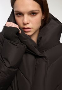 Czarna pikowana kurtka z wysokim kołnierzem. Posiada ściągacze przy mankietach oraz gładką, błyszczącą teksturę.