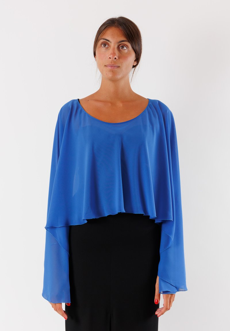 Blusa blu, trasparente, con scollatura arrotondata e maniche lunghe e fluide. L'orlo è irregolare, creando un effetto a strati. Abbinata a un outfit nero.