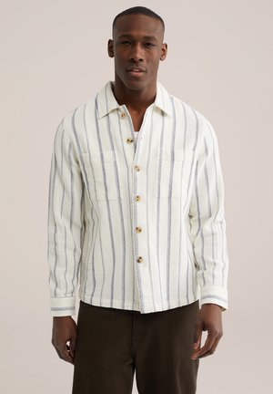 Mies, joka pitää valkoista paitaa, jossa on pystysuuntaiset siniset raidat, ja ruskeat housut, seisoo yksiväristä taustaa vasten.