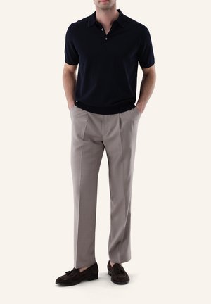 Mann trägt schwarzen kurzärmeligen Poloshirt, beige plissierte Hose und dunkelbraune Loafers mit Quasten, steht mit Händen in den Taschen.