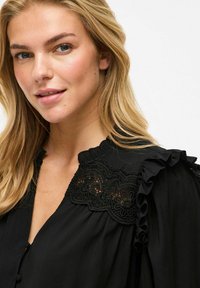 Blouse noire avec des manches à volants et des détails en dentelle intricats sur l'épaule. Tissu doux avec un devant boutonné.