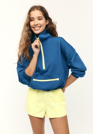 Junge Frau lächelnd, trägt einen blauen Pullover mit gelben Reißverschlüssen und hellgelbe Shorts, posiert mit einer Hand auf der Hüfte und der anderen nahe dem Reißverschluss.