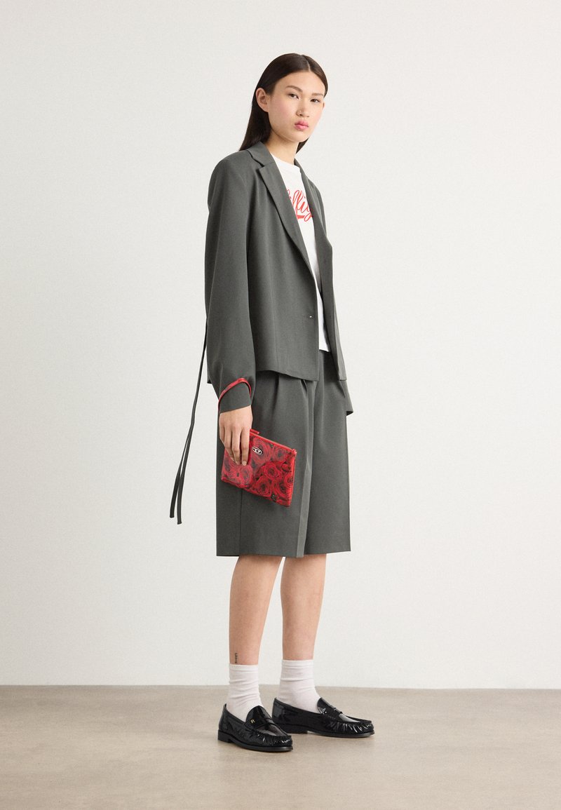Grauer Blazer mit rotem Besatz über einem weißen T-Shirt mit Grafik, kombiniert mit grauen Shorts und schwarzen Slippern. Hält eine rot-blumendekorierte Clutch.