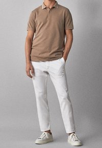 Beżowa koszulka polo z białym kołnierzykiem, w połączeniu z białymi slim-fit spodniami i jasnymi sneakerami. Materiał wydaje się gładki i dobrze dopasowany.