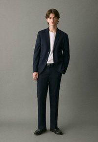 Granatowy garnitur z dopasowaną marynarką i spodniami slim-fit, w połączeniu z białym t-shirtem i czarnymi butami, na neutralnym tle.
