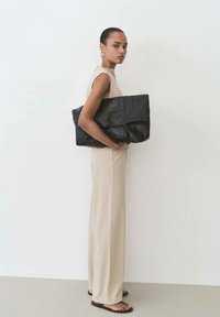 Black leather tote bag with quilted texture, large rectangular shape, and top handles, held by a model wearing a beige jumpsuit.
