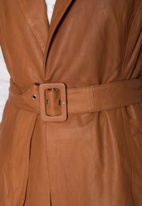 Chaleco de cuero marrón con cinturón, que presenta una hebilla cuadrada, cuello con muescas y detalles de costura, con bolsillos laterales y una textura suave.