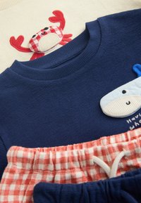 Chemises pour enfants bleu et crème avec appliques cousues de crabe rouge et de baleine bleue, associées à des pantalons à carreaux rouges et blancs avec cordons.