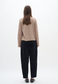 Femme debout de dos, portant un haut marron clair à manches longues, un pantalon noir ample et des chaussures noires sur un fond blanc uni.