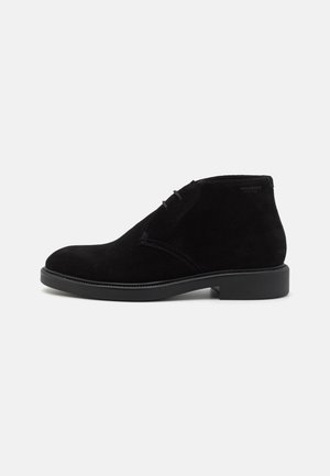 Schwarzer Wildleder-Stiefel mit Schnürung vorne und niedrigem Blockabsatz, Seitenansicht auf einfarbigem hellgrauem Hintergrund.
