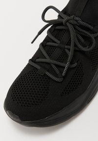 Czarne sportowe buty z siateczkową cholewką, okrągłym czubkiem i teksturowaną podeszwą. Posiadają czarne sznurówki i akcenty oraz gładki, elastyczny design.