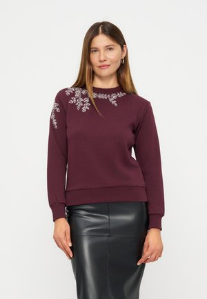 Femme aux cheveux bruns raides portant un sweat-shirt bordeaux avec une broderie florale blanche et une jupe en cuir noire, debout devant un fond blanc.
