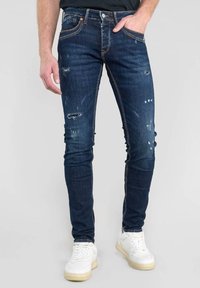 Vaqueros oscuros con detalles desgastados, ajuste slim y costuras naranjas; combinados con zapatillas blancas sobre un fondo neutro.