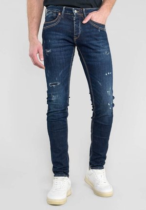 Vaqueros oscuros con detalles desgastados, ajuste slim y costuras naranjas; combinados con zapatillas blancas sobre un fondo neutro.