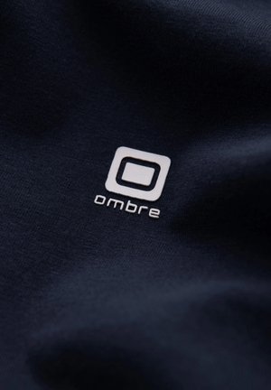 Námořnická tkanina s bílým embosovaným logem "ombre" uvnitř čtvercového tvaru. Hladká textura s minimální pružností, zaměřená na detaily značky.