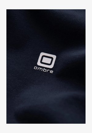 Marine stof met een witte reliëf "ombre" logo binnen een vierkante vorm. Gladde textuur met minimale rek, gericht op merkdetail.