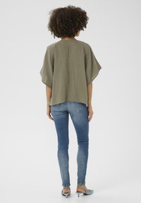 Olíva zöld, laza fazonú felső rövid, denevérujjakkal. Sötétkék, testhez simuló farmernadrággal és világoskék, slip-on sarkú cipővel párosítva. Sima textúrájú anyag.