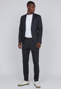 Blazer gris oscuro con corte ajustado y solapas de muesca, combinado con pantalones a juego y una camiseta blanca. El calzado consiste en zapatillas blancas con detalles en verde.