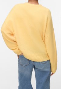 Personne portant un pull jaune ample et doux et un jean bleu en denim, vue de dos avec une main posée sur la hanche.