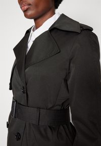 Femme portant un trench-coat noir ceinturé par-dessus une chemise blanche à col, visible du cou jusqu'au milieu du torse sur un fond uni.