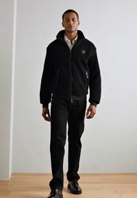 Giacca in fleece nera con zip e colletto alto, tasche laterali e polsini a costine, abbinata a pantaloni neri e scarpe nere lucide.
