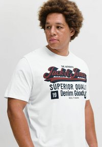 Hvid bomulds t-shirt med et sort og rødt grafisk print; teksten inkluderer "Jack & Jones" og "Superior Quality Denim Goods." Korte ærmer.