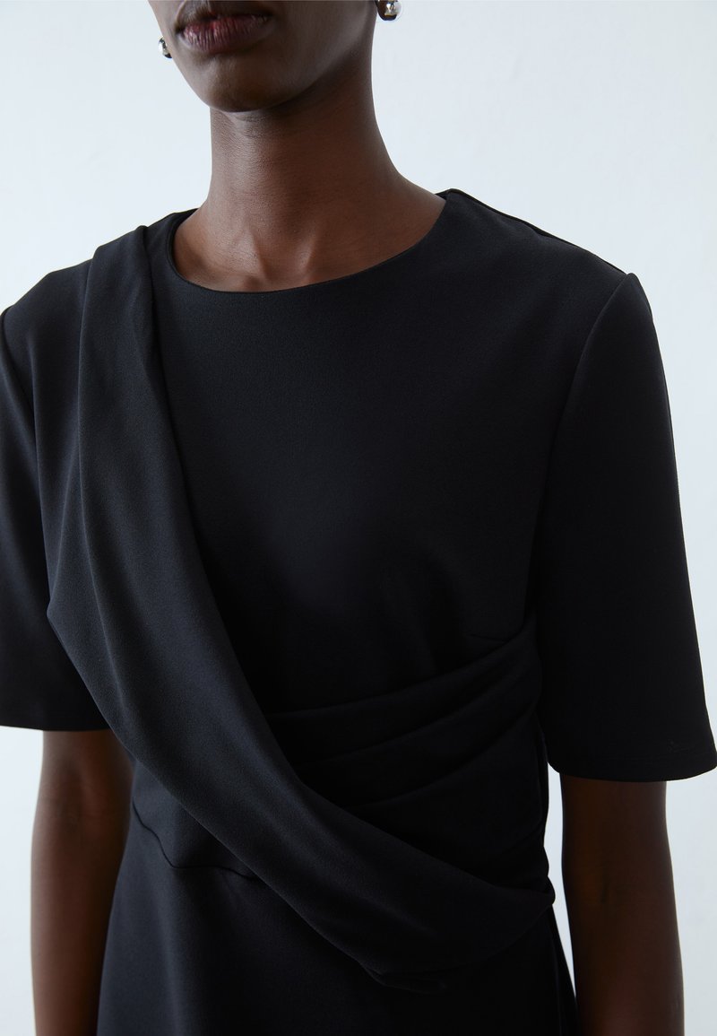 Schwarzes Kleid aus Jersey-Stoff mit kurzen Ärmeln, rundem Ausschnitt und drapierten Details an der Taille. Glatte Textur und figurbetonte Silhouette.