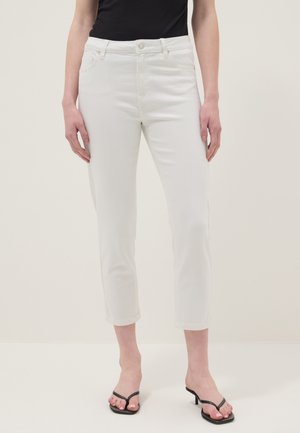 Mujer con jeans blancos recortados, top negro sin mangas y sandalias negras de tacón, de pie frente a un fondo claro y liso.