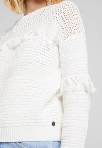 Weißer gestrickter Pullover mit offener Maschenstruktur, Fransenakzenten an den Ärmeln und geripptem Abschluss. Kleines Logo-Detail am Saum.