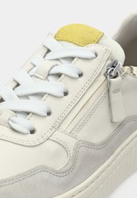 Λευκό αθλητικό παπούτσι από δέρμα με γκρι σουέντ λεπτομέρειες, κίτρινο υφασμάτινο ταμπελάκι, λευκά κορδόνια και πλαϊνό φερμουάρ για εύκολη πρόσβαση. Υφή σε όλη την επιφάνεια.
