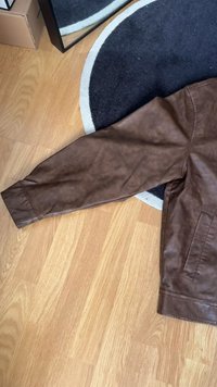 Pruun nahktagi varrukas, millel on sile pind, mansetiga ääris ja küljetasku. Paigutatud puidupõrandale ümmarguse vaiba kõrvale.