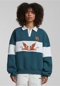 Sweatshirt teal cu o bandă albă în dungi, având un grafic portocaliu cu două creaturi cu aripi și textul "Berlin NY". Design căptușit.