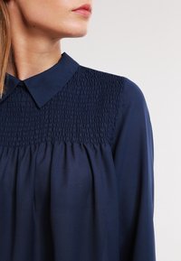 Blusa blu navy con colletto, caratterizzata da una texture arricciata sul petto e un tessuto morbido e fluente che scende dalla vita.