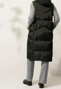 Dlouhá černá péřová vesta s kapucí, se prošívaným designem a šedými pletenými rukávy, doplněná o šedé kalhoty a černé boty.