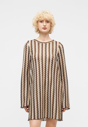 Lyshåret kvinde iført en knælang kjole med lange ærmer og et zigzag-mønster i brunt, beige og hvidt på en lys baggrund.