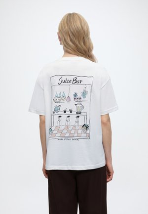 Άτομο με κυματιστό ξανθό μαλλί που φορά λευκό T-shirt με πολύχρωμη εικονογράφηση juice bar, φρούτα, μπλέντερ, σκαμπό και κείμενο στην πλάτη.