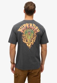 Szary bawełnowy t-shirt z wyrazistą, wielokolorową grafiką smoka otoczoną płomieniami oraz napisem "SUPERDRY" powyżej.