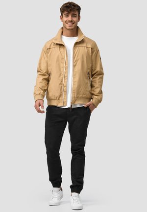 Lysbrun letvæg bomberjakke med lynlås og to sidelommer, parret med sorte joggers og hvide sneakers. Ensfarvet hvid T-shirt underneath.