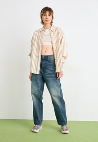 Light beige and striped oversized shirt, cropped white top, dark blue jeans, and patterned sneakers, standing on green floor against white wall.