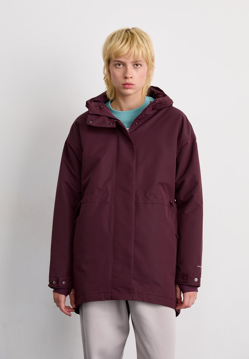 Burgunderfarbige wasserdichte Jacke mit Kapuze, ausgestattet mit Druckknöpfen und Seitentaschen, kombiniert mit einem hellblauen Sweatshirt und grauen Hosen.