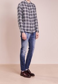 Camicia a righe grigie e bianche con bottoni, jeans chiari strappati blu, e scarpe stringate in pelle marrone scuro, che mostrano uno stile casual moderno.