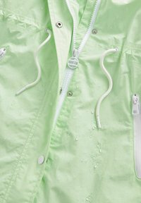 Hellgrüne wasserdichte Jacke mit einem Frontreißverschluss, weißen Akzenten, Kordelzügen und Wassertropfen auf der Oberfläche. Silberne Hardware-Details.