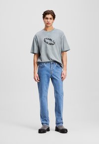 T-shirt gris surdimensionné avec un logo circulaire noir, associé à un jean droit bleu clair et des bottines noires. Design minimaliste, style décontracté.