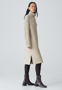 Beiges Strickkleid mit hohem Kragen, seitlichen Schlitzen und strukturiertem Finish, kombiniert mit dunkelbraunen Booties mit Blockabsätzen.