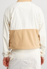 Fleecejacka i kräm och tan, med en strukturerad tyg och en färgblockeringsdesign. Har en elastisk nederkant och justerbara ärmslut.
